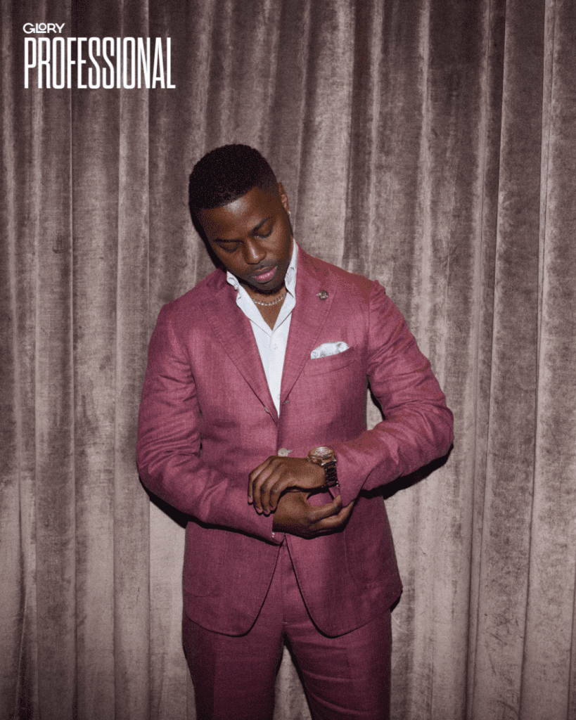 Mean wearing a magenta linen suit and white button up shirt with pocket square. He is looking down at a watch with velvet curtains behind him.