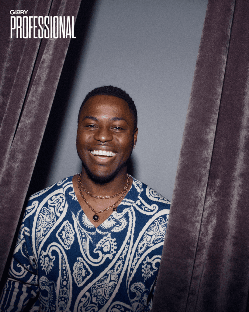 Man wearing a blue paisley sweater smiling inbetween a set of velvet curtains.