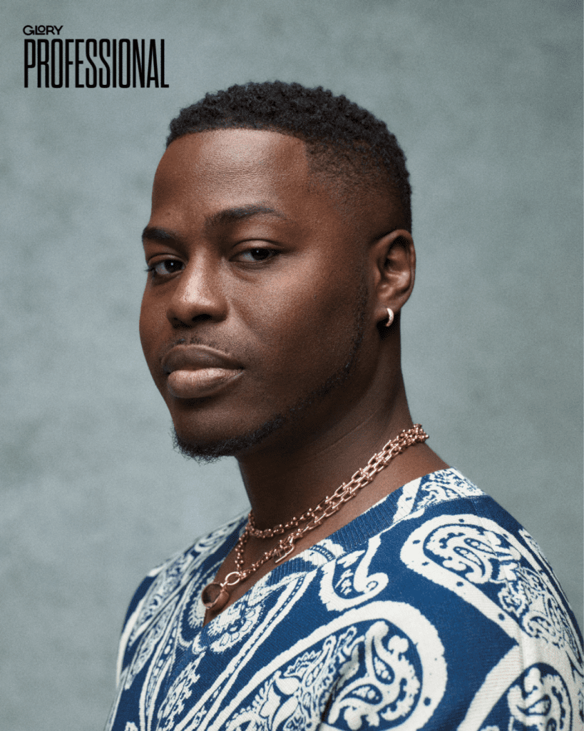 Man wearing blue paisley sweater with big gold jewelry looking into the camera strikingly against a grey background.
