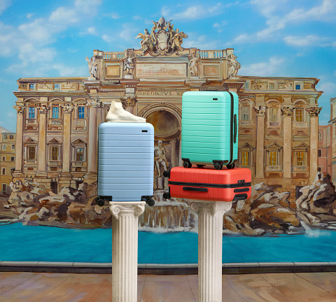 Serena Williams Away Luggage in blue, turquoise, and orange on roman columns against backdrop of fountain Serena Williams Away luggage on columns