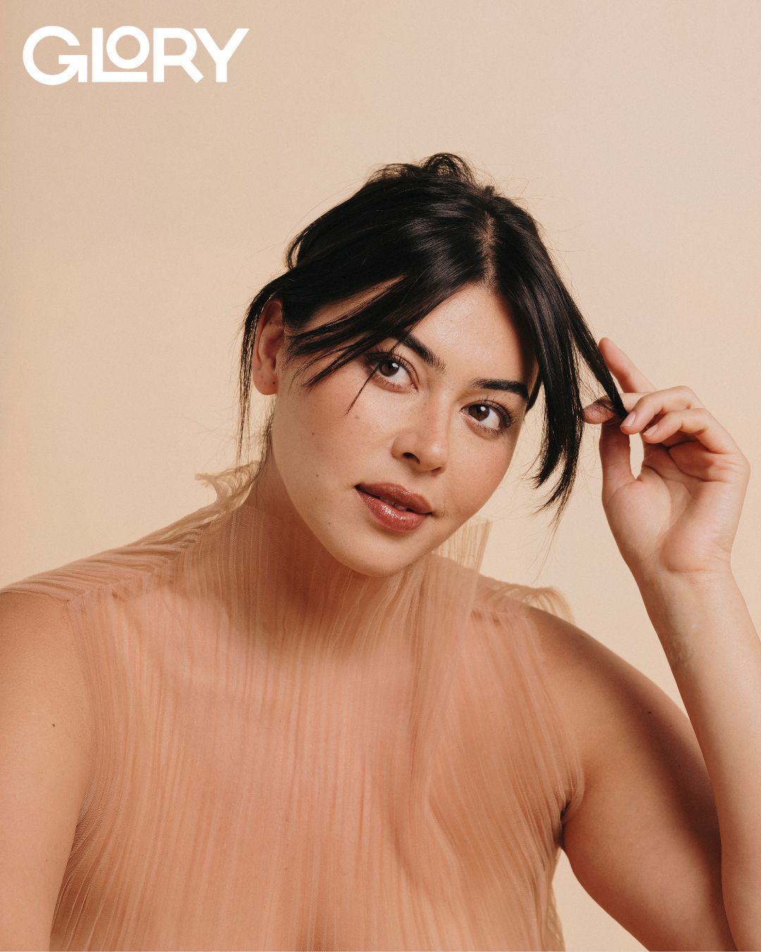 Lauren Chan with her hair tied back. She is wearing a sheer pleated tunic and holding a strand of her hair looking off into the corner.