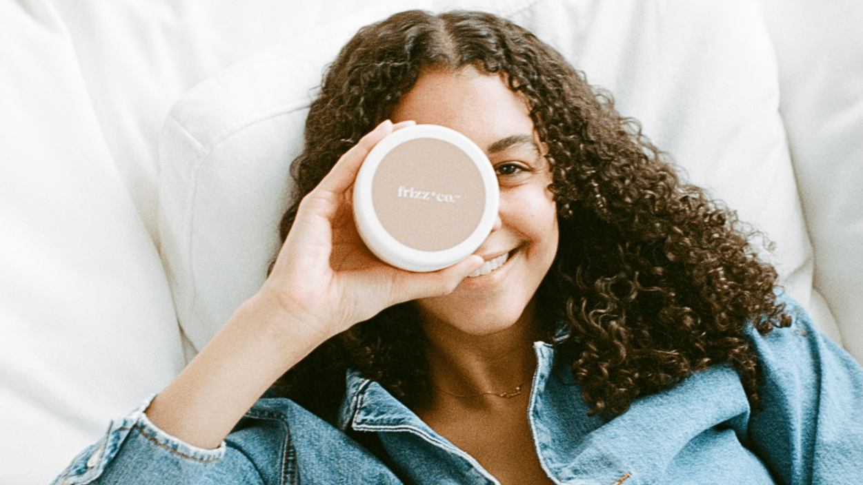 Daphne Eastmon lying on a pillow holding a jar of her Frizz hair cream over her eye.
