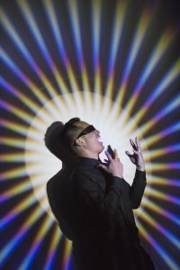 a blindfolded man laughing with colours in a pinwheel formation on the wall behind him