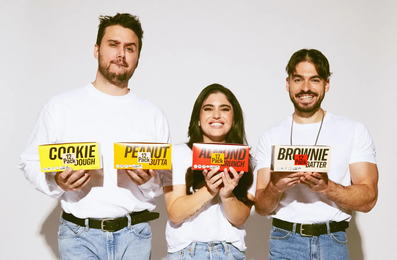Mid-Day Squares founders in white t-shirts and jeans holding their products. There are three of them. Two of them are guys and one is a woman.