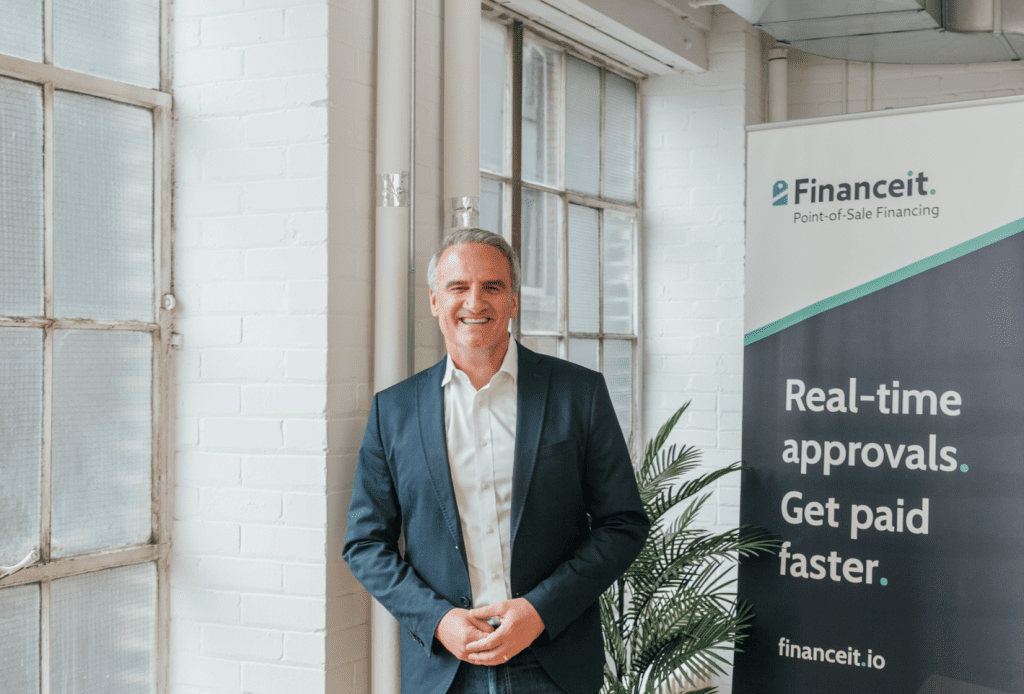 Michael Garrity standing in a room next to a white brick wall and a closed window. He is wearing blue jeans, a white button down shirt, and a navy blazer. On his other side is a plant and a banner that reads "Financeit. Point-of-sale Financing. Real-time approvals. Get paid faster. Fincanceit.io"