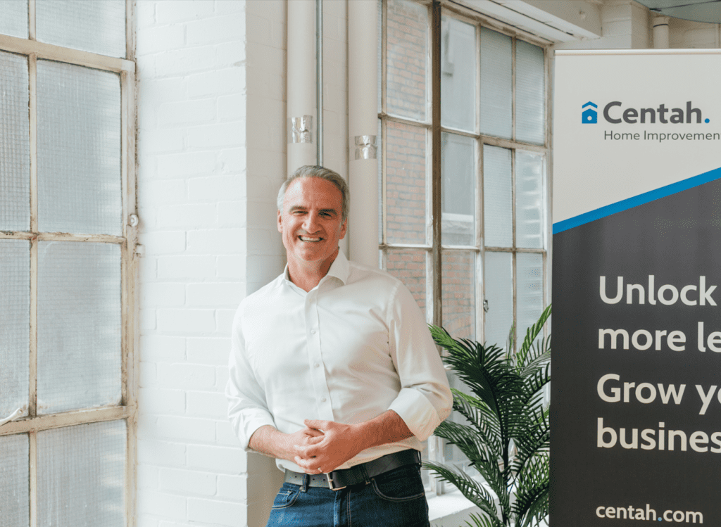 Michael Garrity standing in a room against a white brick wall. On one side of him there is a window, and on the other side there is a plant and a sign. Most of the words on the sign are cut off so it is unclear what it says. Michael is wearing blue jeans and a white button shirt. He is smiling.