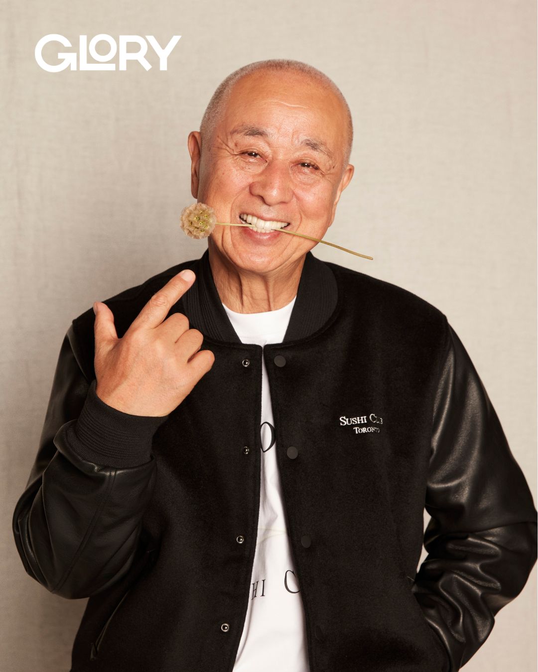 Nobu Matsuhisa wearing a black leather jacket with a dried flower in his mouth. He is pointing at it.