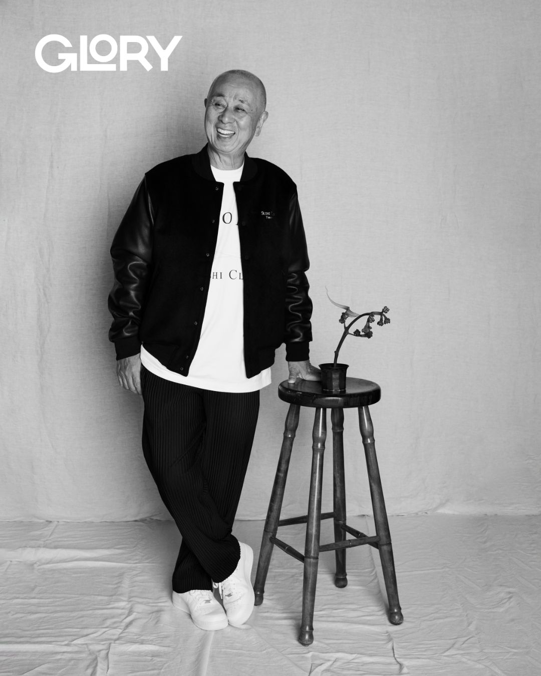 Nobu Matsuhisa wearing a black leather jacket and leaning against a stool with a dried flower arrangement.