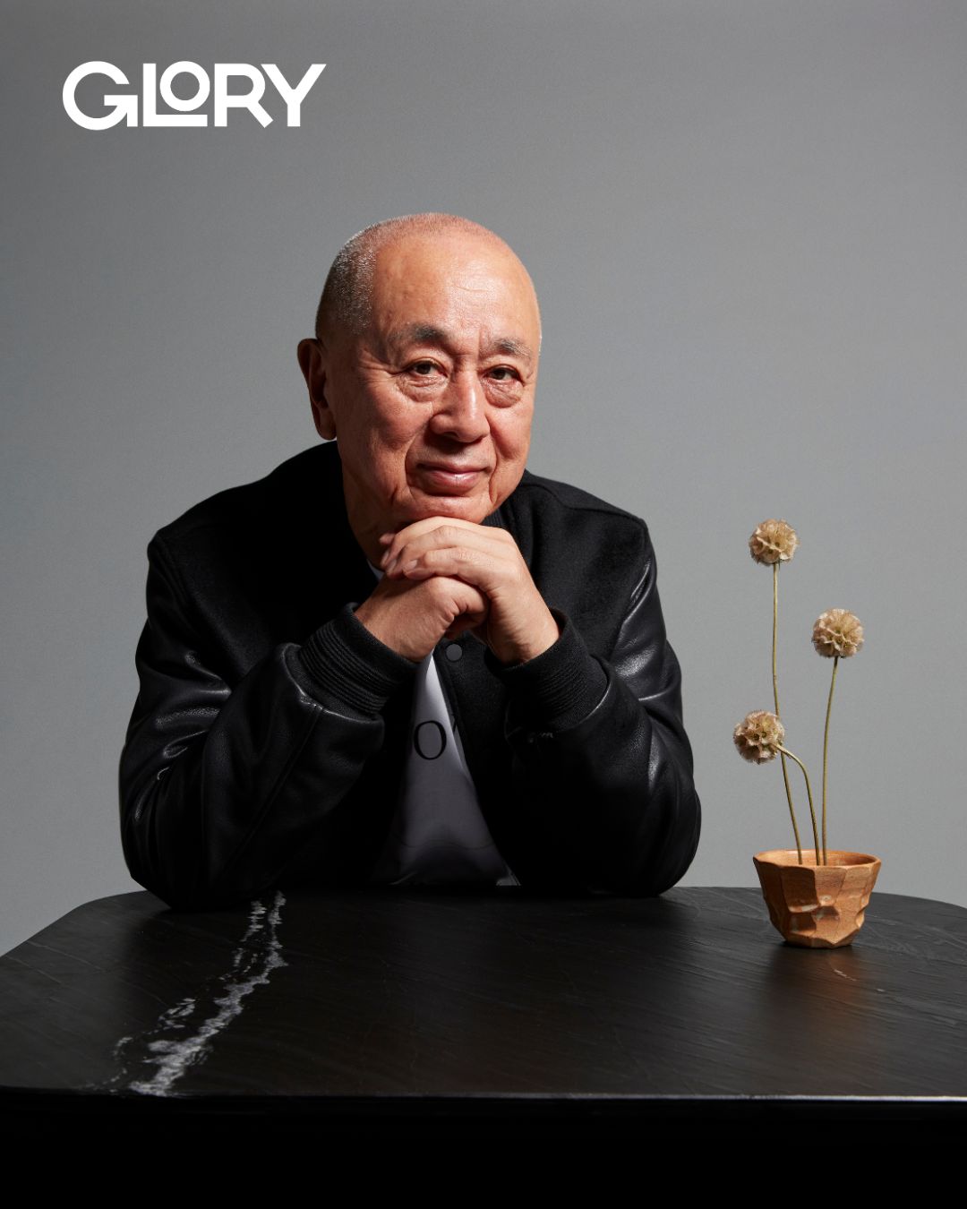 Nobu Matsuhisa wearing a black leather jacket and leaning against a table with a dried flower arrangement.