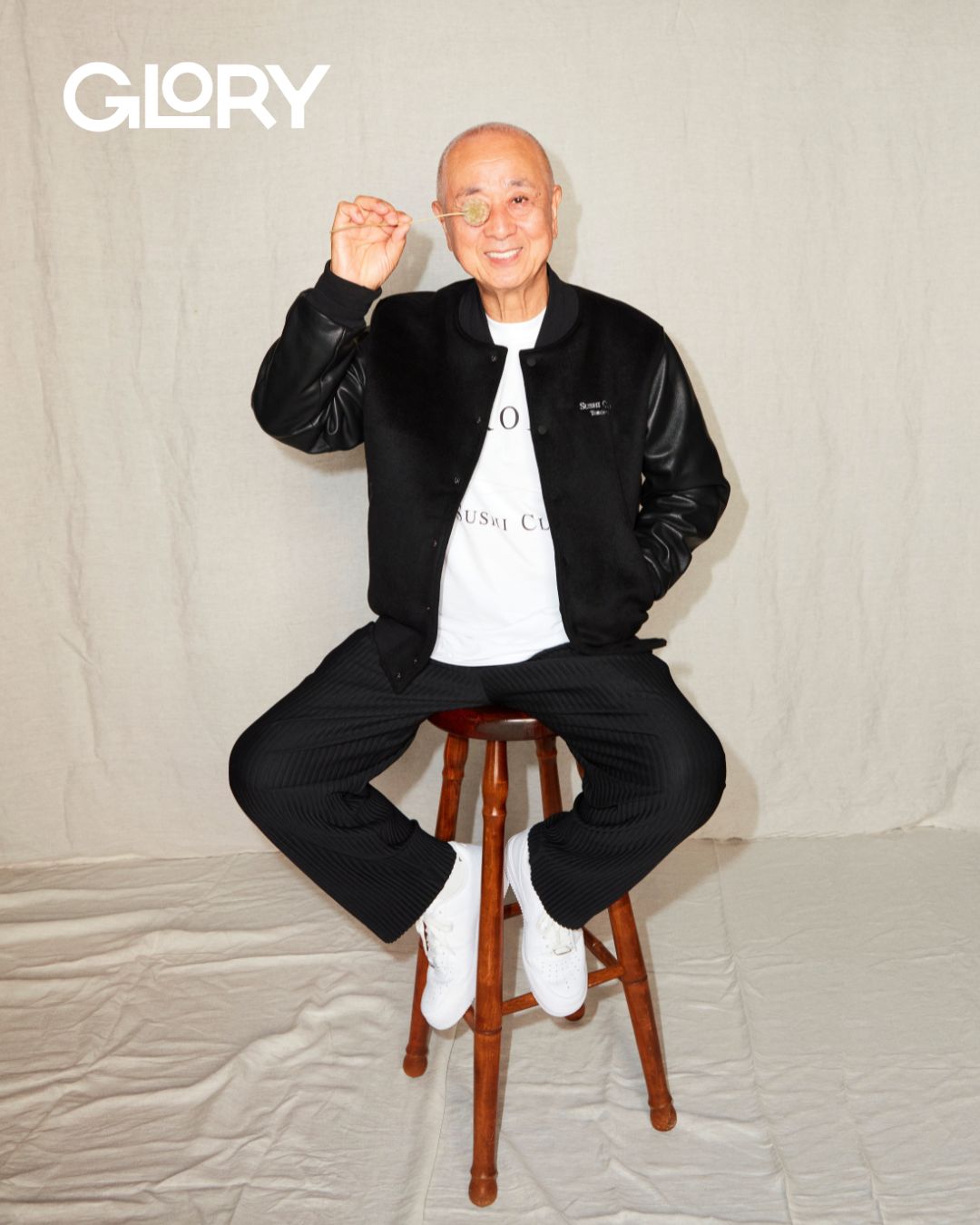 Nobu Matsuhisa wearing a black leather jacket and leaning against a stool with a dried flower arrangement.