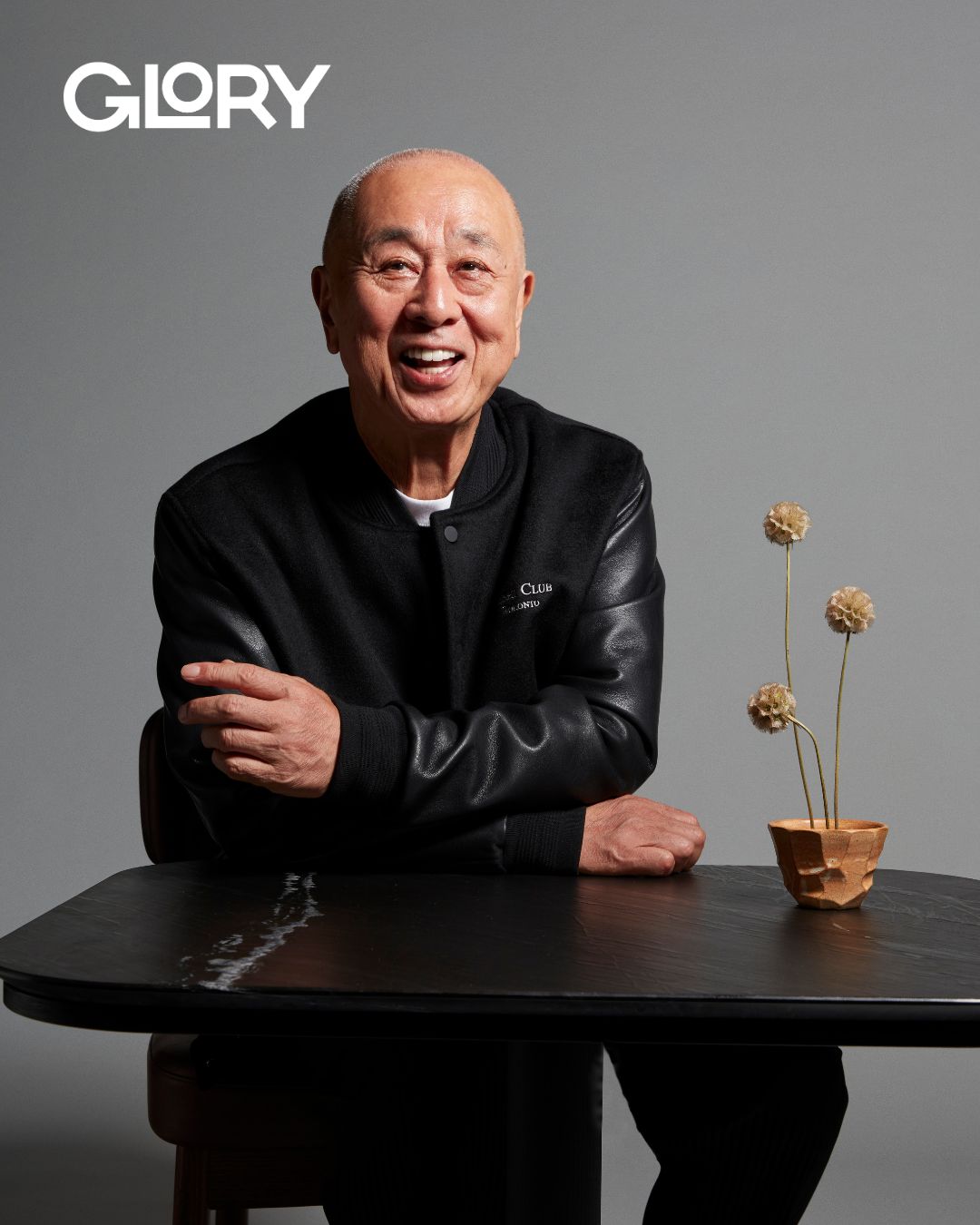 Nobu Matsuhisa wearing a black leather jacket and leaning against a table with a dried flower arrangement.