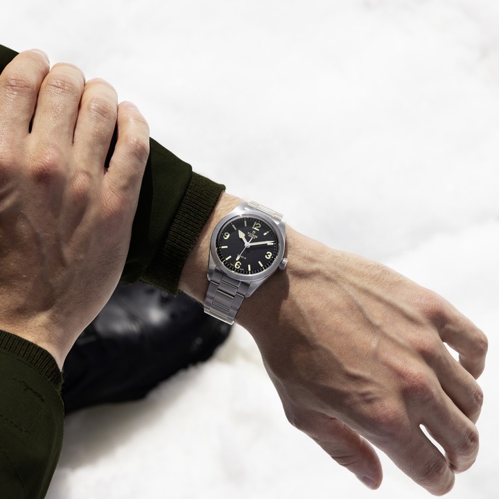 An arm wearing a TUDOR Ranger watch with a steel bracelet. The other hand is grasping the forearm and in the background is a snowbank.