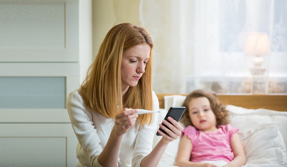 Mother measuring temperature of her ill kid. Sick child with high fever laying in bed and mother holding thermometer. Mother with cell phone calling to doctor