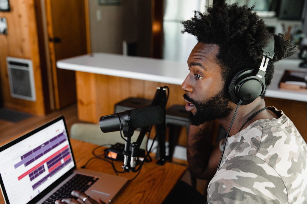 Man wearing a grey camouflage print shirt with headphones speaking into a desktop microphone in front of a wooden desk and a laptop.